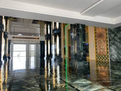 The image shows the interior of a large, modern building, likely a mosque, with a highly polished, dark green marble floor that reflects the lighting and architectural features. The space is characterized by massive, dark marble-clad columns, some of which are illuminated with vertical strips of light. On the right side, there are decorative wall panels with intricate golden Arabic calligraphy and geometric patterns. In the background, a wide opening leads to an area with large windows and natural light, where two figures can be seen, one adult and one child. The overall impression is one of grandeur and opulence. The scene is located in Aluekambuk, Indonesia.