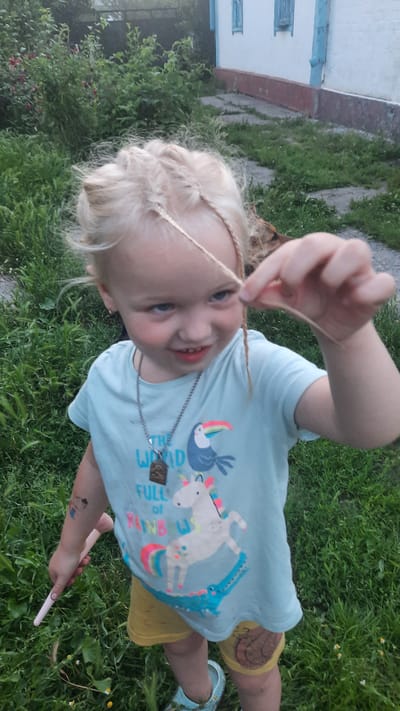 In Samar, Ukraine, a young blonde-haired girl with blue eyes is shown outdoors. She is wearing a light blue t-shirt with a graphic print of a unicorn and a crocodile, along with yellow shorts. The girl has braided sections of her hair and is holding a strand of hair between her fingers, looking at it with a slight smile. She is also wearing a necklace with a pendant and has a light pink object, possibly a wand, in her left hand. The background consists of green grass and some foliage, with a white building with blue trim visible in the distance. The lighting suggests it is daytime.