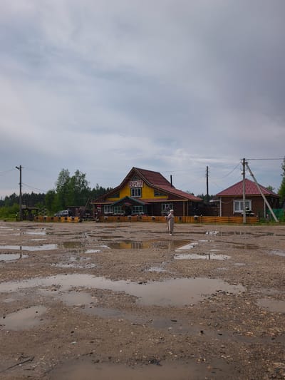 The image depicts an outdoor scene on a cloudy day. The foreground is dominated by a muddy, gravelly ground with numerous large puddles, indicating recent rainfall or poor drainage. In the mid-ground, a cluster of wooden buildings is visible. The largest building, painted yellow with a red roof, has "ВЕРСТА" written in red on its facade. A smaller, darker wooden building with a red roof stands to its right. A single person, dressed in a long, light-colored robe and head covering, stands between the buildings, facing away from the camera. The background features a line of trees and a forest. Electrical poles and wires are present in the scene, suggesting the presence of infrastructure. The overall impression is one of a rural or semi-rural location, possibly a roadside establishment or a small village. Based on the Cyrillic signage, the location is in Russia. Specifically, this scene appears to be at the roadside establishment "Верста" (Versta) located in the village of Kady, Kologrivsky District, Kostroma Oblast, Russia.