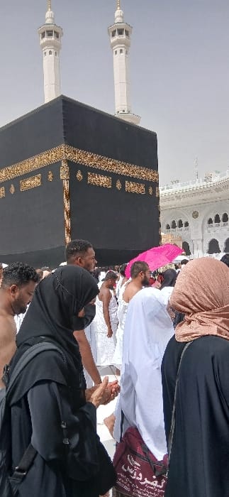 This image depicts a crowded pilgrimage scene around the Kaaba within the Grand Mosque (Masjid al-Haram) in Makkah Al Mukarramah, Saudi Arabia. The central focus is the Kaaba, a large black cuboid structure adorned with gold calligraphic embroidery, occupying the left and central midground. Two white minarets with golden finials are visible against an overcast sky above the Kaaba. Numerous individuals are present in the foreground and midground. Several men are dressed in the white Ihram garments, comprising two unstitched white cloths. Women are predominantly clothed in black abayas and hijabs, with one woman in the right midground wearing a brown hijab. In the immediate foreground, a woman in a black abaya and hijab holds her hands together, palms slightly cupped, in a gesture suggestive of prayer or supplication. To her right, a maroon shoulder bag with white Arabic script is partially visible. In the midground, a male pilgrim holds a bright pink umbrella aloft. White marble architectural structures of the Grand Mosque are visible on the far right. The scene is illuminated by daylight.