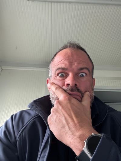 A man is positioned in front of a horizontally ribbed, light-colored wall, possibly a garage door or similar exterior structure. He is wearing a dark-colored jacket, and a smartwatch is visible on his wrist. His face shows an expression of surprise. He has short, thinning gray hair and a salt-and-pepper beard. The man's hand is grasping his own face. Based on visual information alone, there are no details to identify the location as Diekirch, Luxembourg.