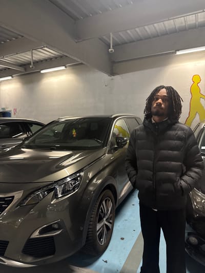 A medium shot depicts a person standing in a parking garage next to a gray car, with the scene presumably located in Montreuil, France. The individual, positioned on the right side of the frame, dons a black puffer jacket and dark pants. The person has dreadlocks. Adjacent to the person is a late-model Peugeot 5008 or similar crossover. The garage environment features a corrugated metal ceiling with fluorescent lighting fixtures. A yellow and red graffiti graphic of a runner can be seen on the wall behind the car. The floor of the parking space is light blue with painted lines.