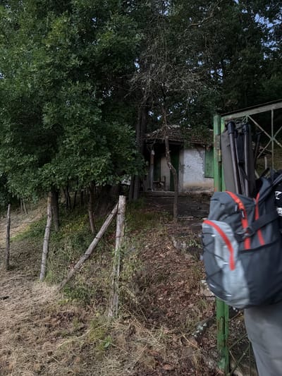A weathered, rustic scene unfolds in Debelets, Bulgaria, near a small, dilapidated house. The house, with its worn facade and tiled roof, sits nestled amongst dense foliage. In the foreground, a person, partially visible, carries a backpack and what appears to be photography equipment, possibly tripods, suggesting an outdoor excursion or work. The surrounding environment is characterized by untamed nature, with overgrown grass, fallen leaves, and rustic wooden fences, hinting at a rural or abandoned setting. The overall impression is one of tranquility, perhaps tinged with a sense of decay, characteristic of less-traveled paths in the Bulgarian countryside.