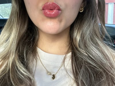 The image shows a close-up of a young woman’s face. She has long, light brown hair that is parted on the side and falls over her shoulders. She is wearing a white t-shirt and gold earrings. Her lips are parted and she is wearing pink lipstick. The woman appears to be in a vehicle. The coordinates (latitude: 25.736, longitude: -100.311) correspond to a location in the city of San Nicolás de los Garza, Mexico.