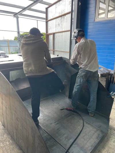 The image shows two individuals working in what appears to be a metal fabrication workshop. The scene is oriented vertically, with the sky at the top and the ground at the bottom.
In the foreground, a person wearing a light-colored hooded jacket and dark pants is bent over, seemingly inspecting or working on a large metal structure. Their face is not visible, but their posture suggests concentration.
Behind them, another person, dressed in a white long-sleeved shirt and blue jeans, is actively welding. They are wearing a welding helmet, which is lifted to reveal their face. A welding torch with a red handle is visible in their hand, and a cable extends from it. The welding sparks are not visible in this particular frame.
The setting includes metal beams and corrugated metal roofing, suggesting an industrial or construction environment. In the background, a blue painted wall and a window with a white frame are visible. The lighting indicates it is daytime, with natural light entering the space. The overall impression is one of active manual labor within a workshop. There is no visible text in the image.