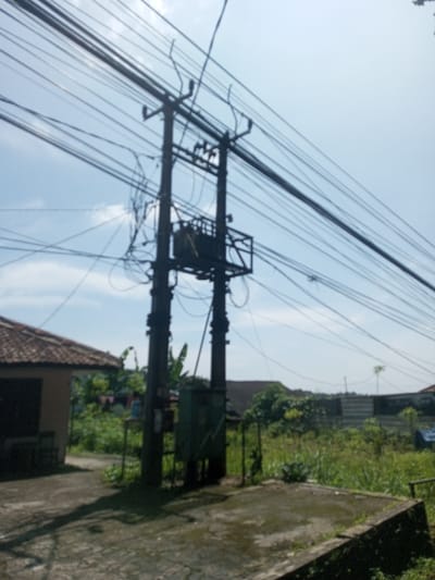 This image depicts an outdoor scene in Beteng_Pangkhar_Somthrang, Bhutan, dominated by electrical infrastructure under a clear sky with scattered clouds. A prominent dark-colored utility pole, possibly concrete or heavily treated wood, stands slightly right of center, comprising two main vertical supports. Mounted high on this pole is a dark electrical transformer, enclosed within a black metal cage structure. Numerous black electrical and communication wires extend horizontally across the upper frame, converging at and emanating from the pole. A tangled mass of wires is visible below the transformer. Near the base of the pole, a green metal electrical distribution box is affixed. The pole rests on a weathered concrete platform or slab that extends into the foreground. To the left, a portion of a building with a reddish-brown tiled roof and light-colored wall is visible, with a small doorway and a dark object resembling a chair or bench outside. The ground in the immediate vicinity of the pole is concrete, exhibiting some discoloration. Beyond the concrete, the landscape transitions into an area of unkempt green grass and foliage. In the background, further structures, vegetation, and a lighter-colored, possibly industrial, barrier are discernible under the bright daylight. No individuals are present in the scene, and no overt actions or interactions are occurring.