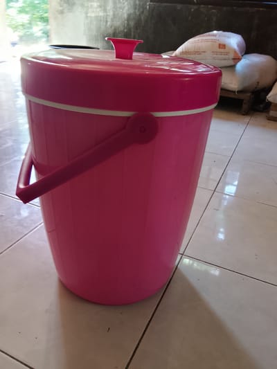 In Merbopuntong, Indonesia, a bright pink plastic container with a lid sits on a tiled floor. The container is cylindrical with a rounded bottom and a handle attached to its side. The lid is also pink and has a small, protruding handle. The floor is made of light-colored square tiles with dark grout lines. In the background, a stack of white sacks is visible, some with text printed on them. The lighting suggests an indoor setting with natural light coming from an unseen source, casting shadows on the floor and the container. The overall impression is one of a utilitarian item in a simple, possibly domestic or storage environment.