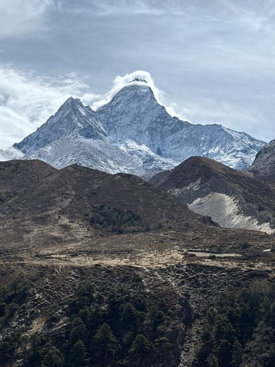 The image presents a grand mountainous landscape, likely captured in the Himalayan region given the context of Khumjung, Nepal. The scene is dominated by two distinct mountain ranges under a partly cloudy, bright sky.
In the background, a colossal, snow-capped peak commands attention. Its rugged surface is extensively covered in brilliant white ice and snow, with swirling clouds partially obscuring its uppermost sections, suggesting high altitude and dynamic weather conditions.
In the midground and foreground, a series of dark, rugged mountains or ridges stretch across the frame. These mountains are composed of dark grey and brown rock, with sparse but visible vegetation. Patches of dark green trees, possibly conifers, and shrub-like growth cling to the slopes, particularly on the right side of the image, providing a contrast to the barren rock. A narrow, lighter-colored path or track is discernible along one of the foreground slopes on the far right, winding its way through the terrain.
The sky is overcast with scattered, wispy clouds, indicating daytime. The light is diffused, suggesting a cloudy day rather than direct sunlight. There are no visible people, animals, or discernible text. The image evokes a sense of vastness, majesty, and the formidable beauty of a high-altitude wilderness.