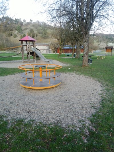 The image displays an unoccupied children's playground situated in the city of Boxberg, Germany. In the immediate foreground, a circular metal merry-go-round with orange railings and a gray platform rests on a light gray gravel surface. Green grass borders the gravel area on the left and right.
In the midground, a wooden playground structure with a red peaked roof and a gray slide is visible. A wooden walkway with chain railings extends to the left from this structure. No individuals are present on or near the playground equipment. To the right, several tall, bare deciduous trees are prominent, with a large tree trunk forming a central vertical element. Behind this tree, a swing set with a dark seat is partially visible. Further right, two wooden picnic tables and a wooden information board with a small peaked roof are situated on a grassy expanse. Two metal trash receptacles are also present near the trees.
The background features a gently sloping hillside covered with mostly bare trees and sparse vegetation, suggesting a late autumn or early spring season. To the left, several red-roofed buildings are discernible. A modern building with scaffolding is partially visible behind the main playground structure on the right. The sky is uniformly bright and overcast, casting diffuse light across the scene.