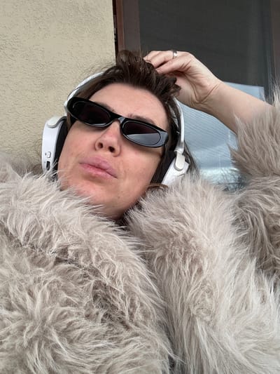 An individual is depicted in a close-up, head and upper torso visible, within what appears to be an outdoor or semi-outdoor setting in Sighnaghi, Georgia. The subject, with dark, disheveled hair, wears small, black, oval-shaped sunglasses and white over-ear headphones. They are dressed in a light grey/beige shaggy faux fur coat. The individual's left hand is positioned with fingers interlocked within their hair, near the top of their head. A silver-colored ring is present on the ring finger of the left hand. The subject's face is directed towards the camera. The background consists of a textured light tan wall on the left and a dark-framed window or doorway on the right, revealing indistinct blurred light blue and grey tones, suggestive of an overcast sky or distant outdoor scene.
