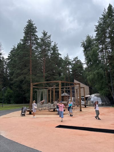 The image depicts an outdoor adventure park or playground in Pavlovskaya Sloboda, Russia. The scene features a complex wooden climbing structure with nets and ropes, designed for challenging activities. Several people are visible interacting with or observing the structure. In the foreground, a woman in a striped dress stands with a bag on her shoulder, looking towards the climbing frame. Further back, a group of individuals, including children and adults, are present near the structure. One person is standing on a higher platform, while others are on the ground level. To the right, a large, opaque, dome-shaped structure is partially visible, possibly a visitor center or unique exhibit. The setting is surrounded by tall, mature pine trees, suggesting a natural, forested environment. The ground surface is a reddish-brown, likely a safety material for the playground. The sky is overcast, indicating a cloudy day. The overall atmosphere appears casual and recreational, with people enjoying an outdoor activity. There is some visible text on a black banner attached to the climbing structure, but it is not legible in this image.