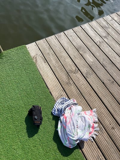 The scene depicts a recreational area adjacent to a body of water in Coruche, Portugal. The image is divided into three distinct zones: a dark, calm body of water in the upper left, a light-colored wooden plank deck running diagonally from the upper right to the lower center, and an area of bright green artificial turf in the lower left. On the artificial turf, a single black watersports glove or wrist support, branded "RADAR" with an orange accent and a central circular fastener, is present. Adjacent to it, partially resting on the wooden deck, is a crumpled pile of textiles. This pile consists of a white garment with black horizontal stripes and black lettering on top, and underneath, a white towel or blanket featuring neon pink and green tie-dye patterns and a fringed edge. Strong overhead sunlight is indicated by the distinct shadows cast by the objects onto the artificial turf and the reflections on the water surface. No individuals are visible in the frame.