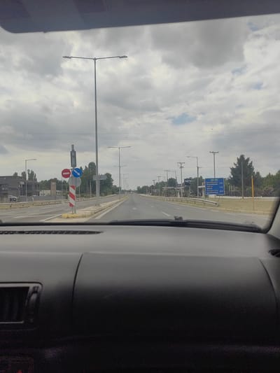 This is a forward-facing, point-of-view shot from inside a vehicle, looking out at a road in Komotini, Greece. The sky is overcast with a mix of white and gray clouds, and a few scattered power lines run horizontally across the upper portion of the image.
The road appears to be a multi-lane highway or a major thoroughfare. On the left side of the road, there are buildings with modern architecture, including a dark gray building with glass windows and a Toyota dealership sign. A car is parked in front of the dealership. Further down the road on the left, there are more buildings and trees.
On the right side of the road, there are streetlights with multiple lamps, interspersed with utility poles. A large blue highway sign is visible, providing directions and distances to several cities: "Θεσσαλονίκη" (Thessaloniki) 242 km, "Αθήνα" (Athens) 734 km, and "Ηγουμενίτσα" (Igoumenitsa) 565 km. Trees and vegetation line the right side of the road.
In the foreground, the black interior of the vehicle, specifically the dashboard, is visible. The dashboard has a textured surface and a vent on the right side.
Traffic signs are present. On the left side of the road, near an intersection or a lane split, there is a pole with several signs. These include a circular red and white sign with a horizontal bar, likely a "do not enter" or similar prohibition, and a blue circular sign with a white arrow pointing to the right, indicating a required turn. A striped red and white barrier is also attached to the pole.