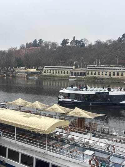 An overcast, high-angle view captures a section of the Vltava River in the Capital City of Prague, Czechia. On the distant, treed hill, identified as Vyšehrad, a prominent red-brick fortification wall is visible on the left, while a building with an ornate, gilded onion dome, recognized as the Church of St. Mary Magdalene, stands on the right. The trees on the hillside appear bare.

Along the river's far bank, a long, light-colored building complex, likely the Výtoň customs house and adjacent structures, features numerous windows and a central columned portico. A chimney on this building emits a plume of white smoke. Behind this complex, several large tour buses are parked. A smaller, modern-looking building is visible further left along the bank, and a small, light-colored dinghy floats in the river near the bank.

In the mid-ground, a dark-hulled passenger boat named "MARIE D'BOHEMIA," operated by "PRAGUE BOATS," navigates the river. Its upper deck is occupied by approximately fifteen individuals, wearing casual clothing and jackets, standing or seated in rows of white chairs. The lower deck is enclosed with large windows.

In the foreground, the upper portion of a larger, stationary passenger boat is visible. Its upper deck is equipped with numerous empty wooden benches and tables. A white canopy, branded "MATTONI," covers the forward section of this deck, while several beige, pyramidal parasols are deployed over the aft section. Multiple orange life preservers are attached to the white railing. No people are visible on this foreground vessel.