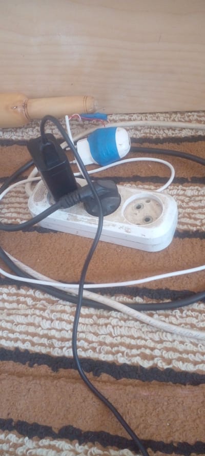 The image shows a multi-plug electrical outlet in what appears to be a domestic setting. The outlet is white and attached to a wooden wall with patterned rug next to it. It has a black plug inserted into one of its sockets. Additional plugs and wires are clustered around the outlet. One plug is white with blue electrical tape. There is a wooden object with what looks like jewelry next to the taped plug.
The location in Parkent, Uzbekistan is not discernable from the image.