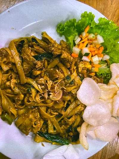 A close-up shot shows a plate of Indonesian food, likely Mie Goreng, served on a white textured plate, placed on a wooden table. The dish features wide, flat noodles coated in a dark, savory sauce, interspersed with pieces of meat and dark green vegetables. Garnishes include a crisp lettuce leaf topped with a diced salad of carrots and cucumbers, and several translucent white crackers (krupuk) arranged on the side. The lighting is warm and direct, creating highlights on the glossy noodles and suggesting an indoor setting, possibly a restaurant or home. The image is framed from a top-down perspective, focusing entirely on the food presentation.