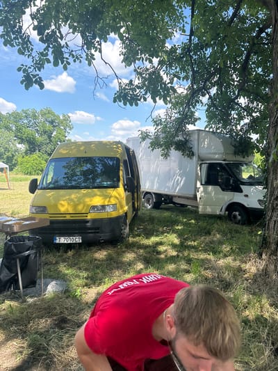 The image depicts an outdoor scene, likely in Lovets, Bulgaria, on a sunny day with scattered clouds. Two vans are parked on a grassy area, partially shaded by a large tree. In the foreground, the back of a person wearing a red t-shirt is visible. The t-shirt has white text on the back, though it is difficult to read fully. A silver chain is visible around the person's neck.

The first van is yellow and faces forward, with its headlights and windshield clearly visible. The license plate reads "T 7010 CT". Next to it, a larger white van is parked, facing the same direction, with its cargo area extending behind the cab. The scene appears to be in a rural or semi-rural setting, with trees and greenery surrounding the vehicles. The sun casts shadows, suggesting it is likely midday. The overall atmosphere seems relaxed and casual, with the person in the red shirt appearing to be part of the scene, perhaps taking a break or observing the surroundings. The presence of a black trash bag on a stand near the yellow van might indicate some form of gathering or activity.