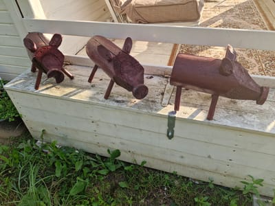 The image shows three wooden piggy decorations, painted a dark reddish-brown, placed on top of a weathered, light-colored wooden storage box. The box is outdoors, situated on a deck or patio area. Green grass and weeds are visible growing around the base of the box. In the background, a white railing and a portion of outdoor furniture with a patterned cushion can be seen, along with a rug on the floor. The scene suggests a rustic, outdoor setting. The image does not provide any specific details about the city of Sorochkino, Russia, nor does it depict any people or actions.
