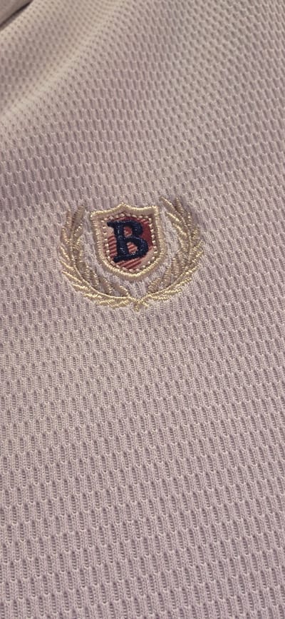 A close-up, eye-level, vertical shot shows a portion of a light purple, mesh-like fabric. Centered in the frame is an embroidered crest featuring a shield with a dark blue letter "B" in the center. The shield is bordered by a thin red and white striped pattern and surrounded by a golden laurel wreath. The fabric is textured with a repeating honeycomb pattern. The image does not contain any contextual information about its location in Collado Villalba, Spain.