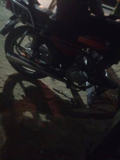 The image shows the rear portion of a motorcycle, likely parked on an uneven surface, potentially dirt or gravel, in Mbribit, Nigeria. The motorcycle is dark in color, with red accents visible on the body panel. The exhaust pipe is prominently displayed. Part of the engine is visible. The image appears to be taken at night or in low light conditions, evidenced by the strong shadows and lack of detail in the background. A person is partially visible on the motorcycle.