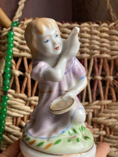 The image shows a close-up, eye-level view of a porcelain figurine of a young girl holding a dove. The figurine is wearing a light pink dress with blue trim. She is kneeling on a green base decorated with orange and green leaf shapes. The background is a woven basket with visible texture and color variations, and a strand of green beads hangs to the left of the figurine. A portion of a hand is visible at the bottom, holding the figurine. The scene appears to be indoors, with diffused lighting. The city of Zelenodol'sk, Russia, is not depicted in this image.