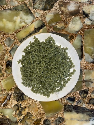 A top-down shot shows a white plate piled high with green pasta on a mottled, brown-and-tan stone surface. The pasta appears to be of the fusilli variety, though with a slightly different, less pronounced spiral. The exact type of stone surface is unknown, but it has a mosaic-like pattern and would appear to be inside a residential location. It is impossible to confirm if the location is in Vicente López, Argentina, based on the visual information provided. There are no people in the image, and the primary subject is the pasta. The action is the presentation of food on a plate. No interactions are visible.