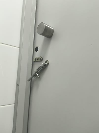 A close-up, eye-level shot shows a light gray metal door with a silver bolt latch and a cylindrical metal handle. The door is ajar, revealing a white tiled wall to the left. The scene is set in a restroom within Vnukovo, Russia. The lighting is even and diffused, suggesting an indoor, possibly public, space. The focus is sharp on the door and its hardware, with the background slightly blurred. No people are present in the frame.
