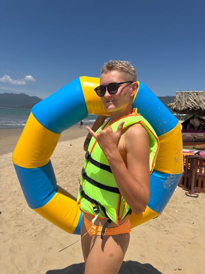 A person wearing a bright green life vest over an orange swimsuit and black sunglasses stands on a sandy beach in Quy Nhon, Vietnam. They are positioned behind a large, inflatable ring that is blue and yellow. The person is holding up a "shaka" hand gesture with their right hand and giving a thumbs up with their left. The background shows the ocean, a coastline with mountains, a thatched-roof hut, and beach chairs. The sky is clear and blue.