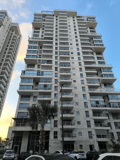 The image depicts a modern residential high-rise building in Ashkelon, Israel, with a second, narrower building to its left. The primary building is light-colored, likely stone or concrete, and features numerous balconies with glass railings. The architecture includes staggered sections and large glass-enclosed areas on multiple floors. A palm tree is prominently positioned in front of the main building's lower levels. Several cars are parked on the street in front of the building, and a lamppost is visible to the left of the main structure. The sky is partly cloudy with hues of orange and yellow from the setting sun visible in the upper left portion of the image. A sign with Hebrew writing is affixed to the building at street level, and another smaller sign with Arabic numerals and Hebrew letters is visible near the building's entrance. The overall scene suggests a contemporary urban residential environment.