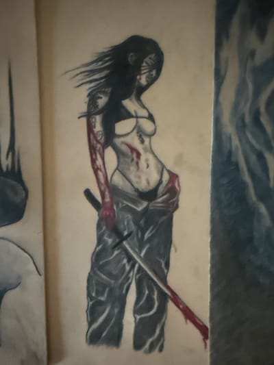 The image depicts a drawing on a wall, featuring a woman with dark hair and what appears to be blood on her arms and face. She is wearing a bikini top and loose-fitting pants, with a sword positioned across her lap. To the left of this drawing, there is a partial depiction of another image, possibly a sculpture or a drawing, with a dark, shadowy form. To the right, a vertical section of the wall displays a blurry, abstract painting with blue and gray tones, resembling flames or water. The overall setting appears to be an indoor space with textured walls, and there are no visible elements that would indicate a specific location in Leominster, United Kingdom.