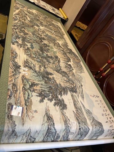 This is a photograph of a large, traditional Chinese ink and wash painting displayed on a green patterned textile. The painting depicts a mountainous landscape with a river winding through it. Several boats are on the river, some with people. Tiny figures of people can be seen walking on a path leading up to buildings nestled on the mountainside. Trees are depicted with fine, detailed brushstrokes, and the mountains are rendered with varying shades of ink to create depth and texture.
In the upper right corner of the painting, there is visible Chinese calligraphy and a red seal. One of the boats in the foreground has a small white tag attached to it with printed text, likely a label or description for the artwork.
The overall scene suggests a serene and tranquil setting, typical of classical Chinese landscape paintings. The use of muted colors and the detailed brushwork evoke a sense of timelessness. The lighting in the photograph suggests it was taken indoors, possibly in a gallery or home, given the wooden ceiling detail visible at the top of the image. The location context provided is Futian District, China.