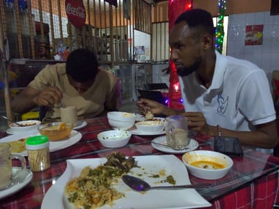 The image depicts two men dining at a casual restaurant, likely during the evening or night, indicated by the dim artificial lighting and colorful string lights visible in the background. They are seated at a table covered with a red and white checkered tablecloth, protected by a clear plastic cover.
On the right, a man with dark skin, a short beard, and close-cropped dark hair wears a white polo shirt with a blue and white logo depicting two stylized figures. He is looking slightly to his left, holding a spoon with food, possibly engaged in conversation or contemplating his meal. Next to his clear glass mug (likely containing chai tea) and saucer, a black smartphone rests on the table.
On the left, another man with dark skin and short dark hair, wearing a light brown or tan short-sleeved shirt, is leaning forward, focused on his food. He is stirring something in a similar clear glass mug with a spoon.
The table is laden with various dishes. In the foreground, a large white rectangular plate holds a partially eaten meal of rice, possibly biryani or pilau, mixed with pieces of meat (which appear to be ribs or stewed pieces) and green herbs, with a spoon resting on it. Several smaller white bowls contain side dishes or sauces, some partially consumed, including one with a yellowish, soupy consistency. A green-capped toothpick holder is also visible on the table.
The background shows a simple restaurant interior with wooden slatted partitions and shelves. A red, oval-shaped Coca-Cola sign with white text hangs prominently. The overall ambiance suggests a relaxed and communal dining experience, typical of establishments in a place like Bagamoyo, Tanzania.