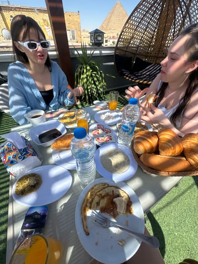 Two women are at a table laden with food and drinks, including croissants in a basket, a plate of pancakes, yogurt cups, and water bottles. One woman is wearing a white tank top and is seated in a wicker chair. The other woman is wearing a blue shirt and white sunglasses. The setting appears to be a rooftop terrace overlooking the city of Giza, Egypt. The Great Pyramid of Giza is visible in the background.