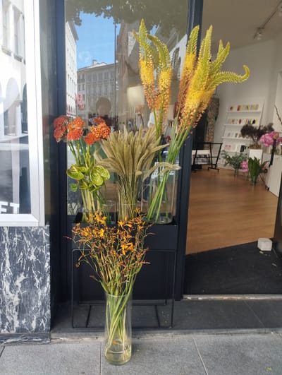A storefront display on a city street features an assortment of flowers. In the foreground, a tall, clear glass vase holds a bouquet of thin, green stems topped with numerous small, orange flowers. To the left of this vase is a black metal stand holding several other floral arrangements. One arrangement consists of tall, thin stalks of dried grass or reeds. Another features bright green, heart-shaped leaves and some red-tipped stems. Behind these, taller, yellow and orange spiky flowers rise from cylindrical glass vases, some of which are partially obscured. The storefront's large glass window reflects the street and buildings across the way, including a person. Inside the store, shelves with various items and additional floral displays are visible. The exterior of the building is characterized by marble-like cladding on the lower portion and a clean, modern window frame. The ground is paved with grey stone slabs. The overall impression is one of a florist's shop presenting its wares outdoors on a sunny day.