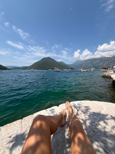 The image shows a first-person perspective of a person's legs and feet resting on a rocky, sun-drenched embankment overlooking a bay in Perast, Montenegro. The person is wearing sandals. The water is a clear, deep blue-green, with gentle waves. In the distance, verdant, tree-covered mountains rise from the water, dotted with buildings. Several boats, including speedboats and a larger, colorful boat with sails, are visible on the bay. The sky is a bright blue with scattered white clouds. The scene captures a serene moment by the sea.
