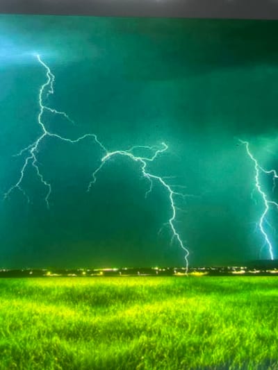 The image depicts a dramatic nighttime thunderstorm scene over the city of Zelenyi Hai, Ukraine. The upper portion of the frame is dominated by a dark, green-hued sky filled with turbulent storm clouds. Multiple lightning strikes are visible; a prominent, intricate bolt descends on the left, while several brighter, more vertical flashes illuminate the clouds on the right. These lightning events cast white to electric blue light across the teal and emerald cloud formations. A thin, horizontal band of bright yellow-white artificial lights, indicative of a distant urban area, stretches across the mid-ground. The lower portion of the image is occupied by an expansive, vividly lit, lime-green field of grass, displaying discernible texture and variations in light consistent with individual blades and clumps. No people, animals, or other specific man-made objects are present within the immediate foreground or mid-ground.