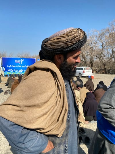 Men, some kneeling on the ground, gather outdoors under a clear sky. A man with a beard and a patterned turban stands in the foreground, wearing a brown shawl over a blue plaid blazer. Other men behind him are dressed in brown and black garments. A white van is visible in the background, along with bare trees. A blue sign with white writing and a graphic is also present.