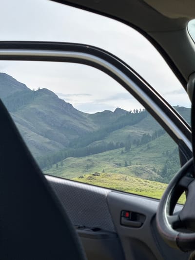 The image shows a view from inside a car, looking out through the open driver's side window at a mountainous landscape. The foreground is dominated by the car's interior, with the edge of a dark grey seat on the left and the steering wheel and door panel on the right. The door panel features a red button and a black door handle. Beyond the window, rolling green hills covered in trees rise towards a cloudy sky. The landscape appears to be rural and undeveloped, characteristic of a natural area like the vicinity of Anchyl-Chon, Russia. No people are visible in the image.