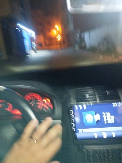 The image presents a blurry, low-light scene from inside a car at night, divided between the vehicle's interior and the external environment.
In the foreground, a human hand, adorned with a simple ring on one finger, rests on a black steering wheel. Behind the steering wheel, the dashboard's instrument cluster glows with red illumination, though specific gauge details are indistinct due to motion blur. To the right, car air vents are visible, along with a digital infotainment screen. This screen, set against a blue background, displays various white, blurred icons and symbols, including one resembling a folder and others suggesting audio or connectivity functions.
Beyond the windshield, a street or alley is illuminated by artificial light sources. On the left, a structure or building emits a prominent blue light. Further down what appears to be a road, several orange streetlights streak across the frame, indicating movement or a long exposure in low light conditions. The overall effect suggests the perspective of being inside a moving or recently stopped vehicle. The time of day is clearly night. No discernible weather conditions are apparent. No legible text is visible within the image. The location context is Martil, Morocco.