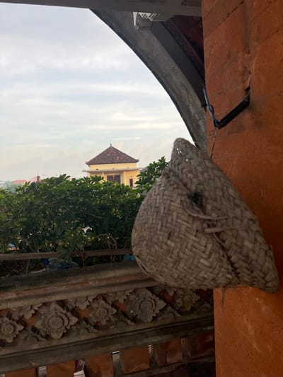 The image shows a close-up, upward view of a traditional woven birdhouse hanging from a red brick structure. In the background, partially obscured by trees, is a building with a red tile roof, characteristic of Indonesian architecture. The sky is overcast. The frame includes elements of a stone wall with floral carvings, typical of the area's artistic style. The location appears to be Celuk, Indonesia, as indicated by the architectural details and regional objects.