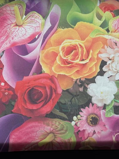 This image depicts a close-up, overhead view of a floral arrangement. The composition is dominated by various flowers in vibrant colors, including a prominent orange rose, a red rose, pink anthurium flowers with green accents, and smaller white and pink flowers. Lush green foliage is interspersed throughout. The arrangement appears to be on a flat surface, possibly a table or as part of a decorative display. There are no people or animals visible in the image, and no context is provided regarding its location within Novoshakhtinsk, Russia. The lighting is even, suggesting an indoor setting or an overcast day.