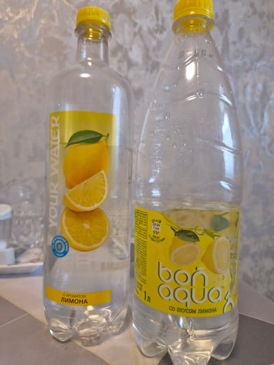The image shows two plastic water bottles with yellow caps. Both bottles are clear and contain water. The top bottle has a yellow label with the text "BonAqua" and "СО ВКУСОМ ЛИМОНА" (with lemon flavor) in Cyrillic. The label also features illustrations of lemons. Below this text, there is nutritional information in Cyrillic. The bottom bottle has a yellow label with "YOUR WATER" in English and "С АРОМАТОМ ЛИМОНА" (with lemon flavor) in Cyrillic. It also has an illustration of lemons and a blue circular seal with Cyrillic text. Both bottles appear to be laid on a surface with a textured pattern, possibly a table. There are some water droplets on the bottles. The lighting suggests it is daytime, possibly indoors. The text on the bottles indicates the product is bottled water with lemon flavor. The location is identified as Zaslawye, Belarus.