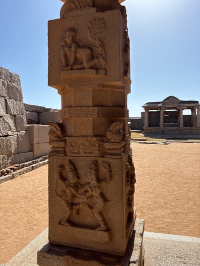 The image shows a detailed stone pillar with carved figures in Kamalapura, India, located in the ruins of Hampi. The pillar is made of a light brown stone and features intricate carvings of human figures in various poses. In the background is a partially destroyed building with multiple levels and pillars, against a clear blue sky. The ground is covered with a layer of sand and stone.