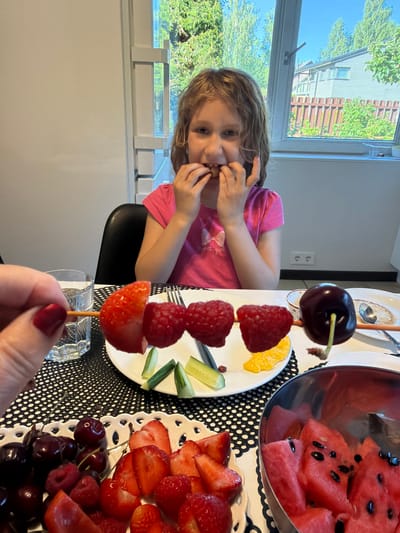 In Baloži, Latvia, a young girl in a pink t-shirt with a bow graphic is seated at a table, eating fruit from a skewer held by an unseen person. The skewer contains a strawberry, raspberries, and a cherry. The table is set with a black and white patterned tablecloth, a glass of water, and plates of various fruits including strawberries, cherries, and watermelon slices in a metal bowl. The background shows a window with a view of trees and houses, suggesting a domestic setting.