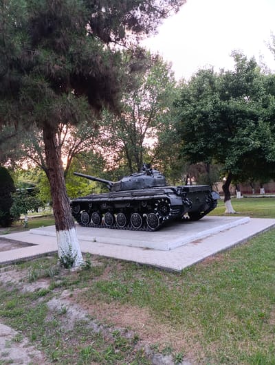 In Fergana City, Uzbekistan, a stationary, dark green military tank is displayed on a raised concrete and paved platform in an outdoor park-like setting. The tank is positioned to face left, with its turret and main gun visible. The platform is bordered by a patterned tiled walkway and surrounded by green grass and mature trees. The trunk of a large tree, painted with white at its base, stands prominently in the foreground to the left, partially obscuring the tank. The background features more trees and a fence, with a pale sky suggesting late afternoon or early morning light. No people are present in the scene.
