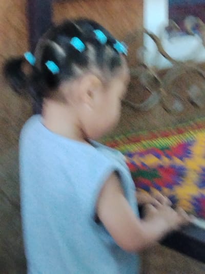 The image shows a young child from the side, with their back to the viewer. The child has dark hair styled in pigtails, adorned with small, bright blue hair accessories. They are wearing a light blue sleeveless shirt. The child's head is tilted down and to the right, with their arm extended forward, appearing to be reaching for something out of frame. The background is blurred, but shows a textured surface that appears to be a woven mat with colorful patterns of red, yellow, green, and blue. To the left of the child, there is a dark vertical object, possibly a door or furniture. The overall lighting suggests an indoor setting, possibly during the day. The image quality is low, with significant blur and grain, making it difficult to discern specific details of the child's face or the exact nature of their activity. The setting and the child's hairstyle suggest a possible cultural context, perhaps related to Ala-Buka, Kyrgyzstan.