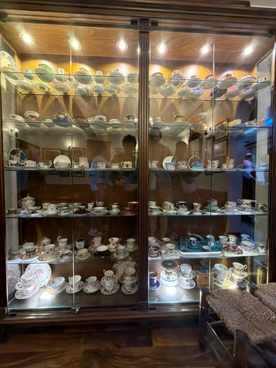 The image shows a glass-fronted display cabinet filled with a collection of antique teacups and saucers. The cabinet has multiple shelves, each displaying various sets of porcelain tableware, some with intricate floral patterns and gold accents. The lighting within the cabinet highlights the delicate craftsmanship of the pieces. The cabinet itself appears to be made of dark wood, with a polished finish. Reflected in the glass are glimpses of the surrounding environment, including wooden walls, framed pictures, and what appears to be a person holding a mobile phone, suggesting the collection is housed in a museum or private collection, possibly in Eyüpsultan, Turkey, a district known for its historical and cultural sites, which often include such displays. The overall impression is one of a curated and well-preserved collection of decorative ceramic ware.
