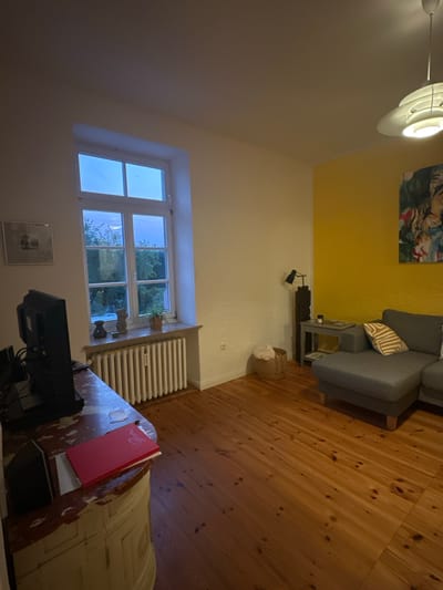 The image shows a living room in Ammeldingen bei Neuerburg, Germany. A large window with a white frame overlooks trees and the sky. Below the window, a white radiator runs across the wall. To the left of the window is a framed picture on the wall, and a television set sits on a distressed wooden cabinet. A red book lies on the cabinet. To the right of the window, the wall is painted bright yellow and features a modern abstract painting. A grey sectional sofa is positioned against the yellow wall, adorned with a striped pillow. A small side table next to the sofa holds a lamp, a candle, and some magazines. A wicker basket filled with blankets sits on the wooden floor near the side table. The floor is made of wooden planks. A ceiling lamp with multiple tiers is visible in the upper right corner of the image. The room appears to be dimly lit, suggesting it is either dusk or nighttime.