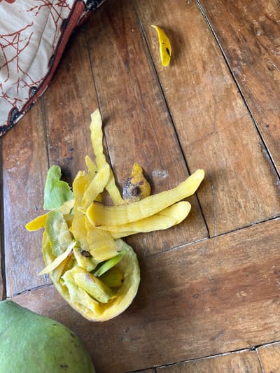An overhead shot of a wooden table in Benuaraja, Indonesia, shows the remnants of peeled mangoes. In the center, a hollowed-out mango skin is filled with mango flesh and peelings. Scattered around it are more mango peelings, some of which are yellow and some green. A single, small piece of yellow peel lies separate from the main pile, to the top right of the image. To the bottom left, the curved side of an unpeeled green mango is visible. In the top left corner, a patterned cloth with red and white designs is partially visible. The lighting is natural, casting subtle shadows.