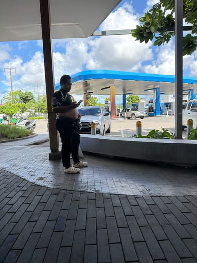 A man with dark hair is standing on a tiled walkway and looking at his phone. He is wearing a black t-shirt with a colorful design on the sleeves and chest, black pants, and white Croc-style shoes. He has a visible belly. Behind him, there is a gas station with blue awnings and pumps. Several vehicles, including a silver truck and motorcycles, are visible at the gas station. In the background, there are trees and a cloudy sky. The man appears to be at a gas station in Si Sunthon, Thailand.